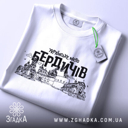 Футболка місто Бердичів, біла, зображена зверху, складена на поверхні, фон світлий, принт містить чорний контурний малюнок архітектурних пам'яток Бердичева з написом 'УКРАЇНСЬКЕ МІСТО БЕРДИЧІВ', на футболці є етикетка з зеленим шнурком.