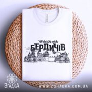 Футболка місто Бердичів, біла, зображена зверху, складена на плетеній основі, фон натуральний, принт містить чорний контурний малюнок архітектурних пам'яток Бердичева з написом 'УКРАЇНСЬКЕ МІСТО БЕРДИЧІВ'.