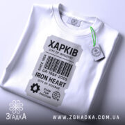 Футболка місто Харків, біла, розкладена на поверхні, фон світло-сірий, принт у вигляді етикетки з написом 'ХАРКІВ', прикріплена бірка зеленого кольору, що додає акценту до дизайну.