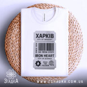 Футболка місто Харків, біла, розкладена на плетеній кошику, фон нейтральний, принт у вигляді етикетки з написом 'ХАРКІВ', оформлення додає естетики, підкреслюючи стиль футболки.