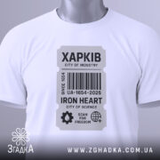 Футболка місто Харків, біла, розкладена на поверхні, фон світло-сірий, принт у вигляді етикетки з написом 'ХАРКІВ', прикріплена бірка зеленого кольору, що додає акценту до дизайну.