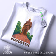 Футболка місто Коростень, біла, складена на столі, фон світло-сірий, принт з зображенням воїна на кам'яній основі, етикетка з зеленим шнурком.