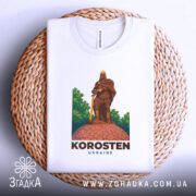 Футболка місто Коростень, біла, складена на плетеній основі, фон світло-сірий, принт з зображенням воїна на кам'яній основі, етикетка видима.