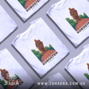 Футболка місто Коростень, біла, зображена на манекені з правого боку, фон світло-сірий, принт з зображенням воїна на кам'яній основі з зеленим фоном.