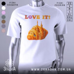 Футболка місто круасан, біла, зображена на манекені, фон світло-сірий, принт з оранжевим круасаном і архітектурними елементами, текст 'LOVE IT!' у кольорах від рожевого до оранжевого.