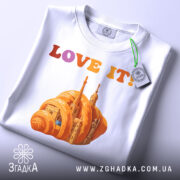 Футболка місто круасан, біла, складена на поверхні, фон світло-сірий, принт з оранжевим круасаном і архітектурними елементами, текст 'LOVE IT!' у кольорах від рожевого до оранжевого, з етикеткою зеленого кольору.