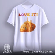 Футболка місто круасан, біла, підвішена на вішалці, фон світло-сірий, принт з оранжевим круасаном і архітектурними елементами, текст 'LOVE IT!' у кольорах від рожевого до оранжевого.