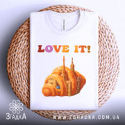 Футболка місто круасан, біла, складена на плетеній кошику, фон світло-сірий, принт з оранжевим круасаном і архітектурними елементами, текст 'LOVE IT!' у кольорах від рожевого до оранжевого.