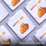 Футболка місто круасан, біла, зображена на манекені, фон світло-сірий, принт з оранжевим круасаном і архітектурними елементами, текст 'LOVE IT!' у кольорах від рожевого до оранжевого.