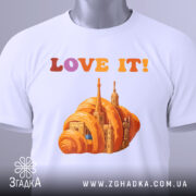 Футболка місто круасан, біла, складена на поверхні, фон світло-сірий, принт з оранжевим круасаном і архітектурними елементами, текст 'LOVE IT!' у кольорах від рожевого до оранжевого, з етикеткою зеленого кольору.