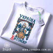 Футболка місто Житомир, біла, розкладена на поверхні, фон світло-сірий, принт з дівчиною, сокирою та левом, текст 'УКРАЇНА ЖИТОМІР' у кольорах синього та червоного, на футболці є етикетка з зеленим шнурком.