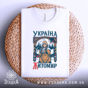 Футболка місто Житомир, біла, розкладена на плетеній кошику, фон світло-сірий, принт з дівчиною, сокирою та левом, текст 'УКРАЇНА ЖИТОМІР' у кольорах синього та червоного.