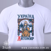 Футболка місто Житомир, біла, розкладена на поверхні, фон світло-сірий, принт з дівчиною, сокирою та левом, текст 'УКРАЇНА ЖИТОМІР' у кольорах синього та червоного, на футболці є етикетка з зеленим шнурком.