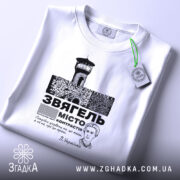 Футболка місто Звягель, білого кольору, складена на поверхні, фон світлий, принт зображає замок та текст, прикріплена бірка з зеленим шнурком.