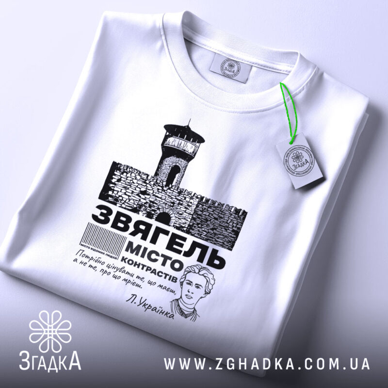 Футболка місто Звягель, білого кольору, складена на поверхні, фон світлий, принт зображає замок та текст, прикріплена бірка з зеленим шнурком.