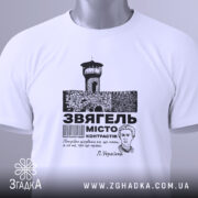 Футболка місто Звягель, білого кольору, складена на поверхні, фон світлий, принт зображає замок та текст, прикріплена бірка з зеленим шнурком.