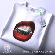 Футболка модна провокація, біла, складена на столі, фон світло-сірий, принт з червоними губами та золотими зубами, на футболці є етикетка з зеленим шнурком.