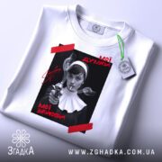 Футболка мої думки зайоби. Біла футболка, складена на столі, з принтом на чорному фоні, де зображена жінка з пістолетом. На принті червоні написи 'Мої думки' та 'Мої зайоби'. На футболці зелений ярлик, фон світло-сірий.