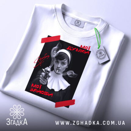 Футболка мої думки зайоби. Біла футболка, складена на столі, з принтом на чорному фоні, де зображена жінка з пістолетом. На принті червоні написи 'Мої думки' та 'Мої зайоби'. На футболці зелений ярлик, фон світло-сірий.
