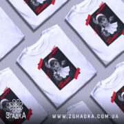 Футболка мої думки зайоби. Біла футболка на манекені, з принтом на чорному фоні, де зображена жінка в чорному вбранні з пістолетом. На принті червоні написи 'Мої думки' та 'Мої зайоби'. Фон світло-сірий, футболка має короткі рукави.