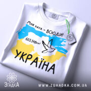 Футболка моя хата всюди, біла футболка, складена на столі, фон світлий, принт з картою України, жовто-блакитні кольори, напис '603.548км²' та малюнок голуба, на футболці є етикетка.