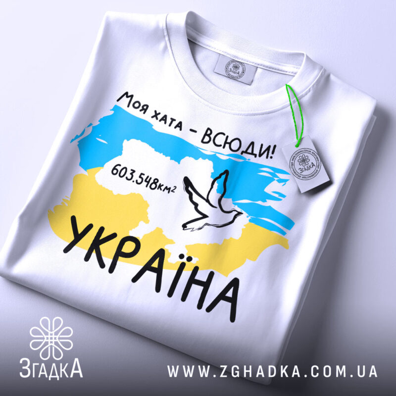 Футболка моя хата всюди, біла футболка, складена на столі, фон світлий, принт з картою України, жовто-блакитні кольори, напис '603.548км²' та малюнок голуба, на футболці є етикетка.