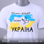 Футболка моя хата всюди, біла футболка, складена на столі, фон світлий, принт з картою України, жовто-блакитні кольори, напис '603.548км²' та малюнок голуба, на футболці є етикетка.
