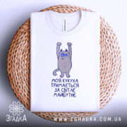 Футболка моя кукуха тримається на плетеній основі, білого кольору, з принтом на грудях, зображаючим персонажа з піднятими руками, фон світлий.