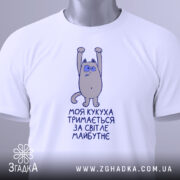 Футболка моя кукуха тримається на столі, білого кольору, з принтом на грудях, зображаючим персонажа з піднятими руками, на комірці є етикетка, фон світлий.