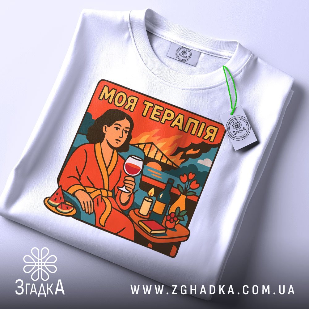 Футболка моя терапія. Біла футболка, складена на столі, з принтом жінки в червоному халаті, що тримає келих з вином. Принт яскравий, з деталями, такими як квіти та свічка.
