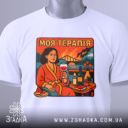 Футболка моя терапія. Біла футболка, складена на столі, з принтом жінки в червоному халаті, що тримає келих з вином. Принт яскравий, з деталями, такими як квіти та свічка.