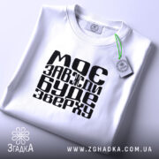 Футболка моє буде зверху, біла, зображена з верхнього ракурсу, на світлому фоні, складена з чорним принтом, що містить текст "МОЄ ЗАВЖДИ БУДЕ ЗВЕРХУ", з етикеткою, що звисає.