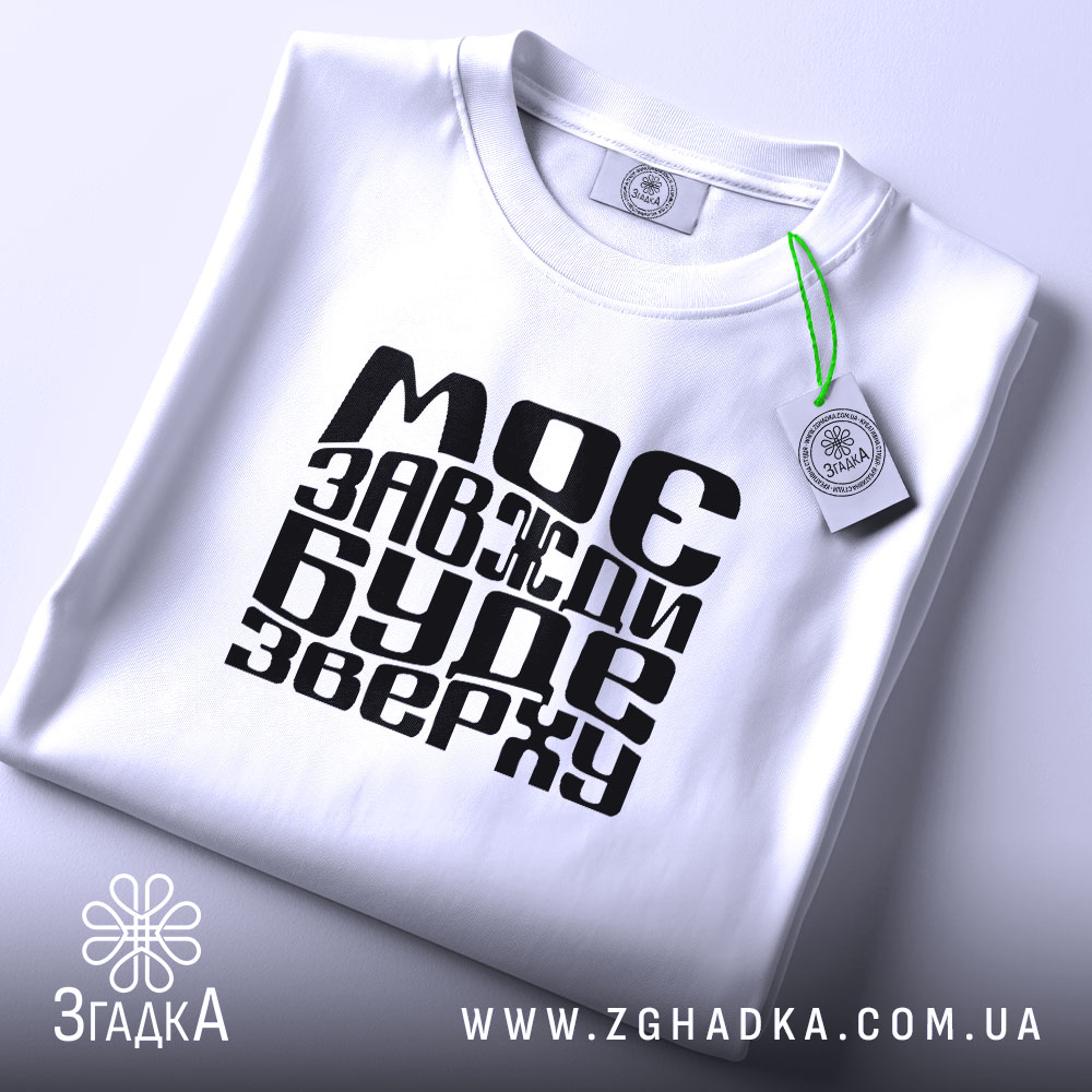Футболка моє буде зверху, біла, зображена з верхнього ракурсу, на світлому фоні, складена з чорним принтом, що містить текст "МОЄ ЗАВЖДИ БУДЕ ЗВЕРХУ", з етикеткою, що звисає.