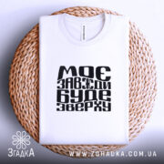 Футболка моє буде зверху, біла, зображена з верхнього ракурсу, на плетеній основі, з чорним принтом, що містить текст "МОЄ ЗАВЖДИ БУДЕ ЗВЕРХУ", складена акуратно.