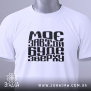 Футболка моє буде зверху, біла, зображена з верхнього ракурсу, на світлому фоні, складена з чорним принтом, що містить текст "МОЄ ЗАВЖДИ БУДЕ ЗВЕРХУ", з етикеткою, що звисає.