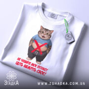 Футболка моє пузіко, біла, зображена зверху на столі, фон світло-сірий, принт з котом у синій футболці, текст червоного кольору, на футболці є етикетка з зеленим шнурком.