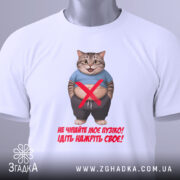 Футболка моє пузіко, біла, зображена зверху на столі, фон світло-сірий, принт з котом у синій футболці, текст червоного кольору, на футболці є етикетка з зеленим шнурком.