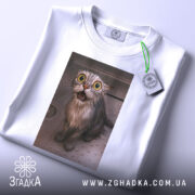 Футболка мокрий кіт, біла, зображення мокрого кота з великими очима на принті, ракурс зверху, футболка складена, фон світлий, присутня етикетка з зеленим шнурком.