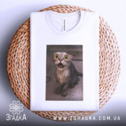 Футболка мокрий кіт, біла, зображення мокрого кота з великими очима на принті, ракурс зверху, футболка складена на плетеній основі, фон світлий.