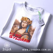 Футболка Мона Будьмо білого кольору, зображена зверху на плоскій поверхні, фон світло-сірий. Принт з жінкою у вінку з квітів, яка тримає стакан, з написом 'Ну що, Будьмо!' червоним кольором, на футболці також видно етикетку.