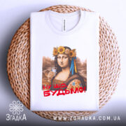 Футболка Мона Будьмо білого кольору, зображена зверху на плоскій поверхні, фон світло-сірий. Принт з жінкою у вінку з квітів, яка тримає стакан, з написом 'Ну що, Будьмо!' червоним кольором, футболка лежить на плетеній основі.