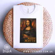 Футболка Мона Ліза дрон білого кольору, зображена зверху на плетеному кошику, фон світлий, принт з Моною Лізою, яка тримає дрон, підпис "ЖИВИ ЖИТТЯ" внизу.