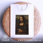 Футболка Мона Ліза білого кольору, лежить на плетеній кошику, фон світло-сірий, принт з портретом Мона Лізи з написом 'Готична стабільність'.