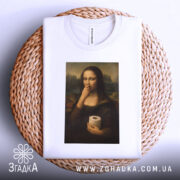 Футболка Мона мем, біла, розкладена на плетеній кошику, фон світло-сірий, принт з Моною Лізою, яка тримає рулон туалетного паперу, вид зверху.