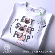 Футболка мопс ритуал, біла, складена на поверхні, фон світло-сірий, принт з текстом 'EAT SLEEP POOP (REPEAT)' та малюнками мопсів, на футболці є етикетка з зеленим шнурком.
