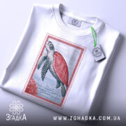 Футболка морська черепаха, біла, складена на столі, фон світло-сірий, принт з морською черепахою в червоній рамці, з етикеткою зеленого кольору.