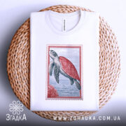 Футболка морська черепаха, біла, складена на плетеній кошику, фон світло-сірий, принт з морською черепахою в червоній рамці, з видимими деталями принта.