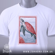 Футболка морська черепаха, біла, складена на столі, фон світло-сірий, принт з морською черепахою в червоній рамці, з етикеткою зеленого кольору.