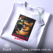 Футболка мотаюсь по бізнесу, білого кольору, розкладена на поверхні, фон світлий, принт з коргі в окулярах, що сидить за кермом, з текстом "Мотаюсь по бізнесу!" червоного кольору, на футболці є етикетка з зеленим шнурком.