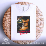 Футболка мотаюсь по бізнесу, білого кольору, складена на плетеній кошику, фон світлий, принт з коргі в окулярах, що сидить за кермом, з текстом "Мотаюсь по бізнесу!" червоного кольору.